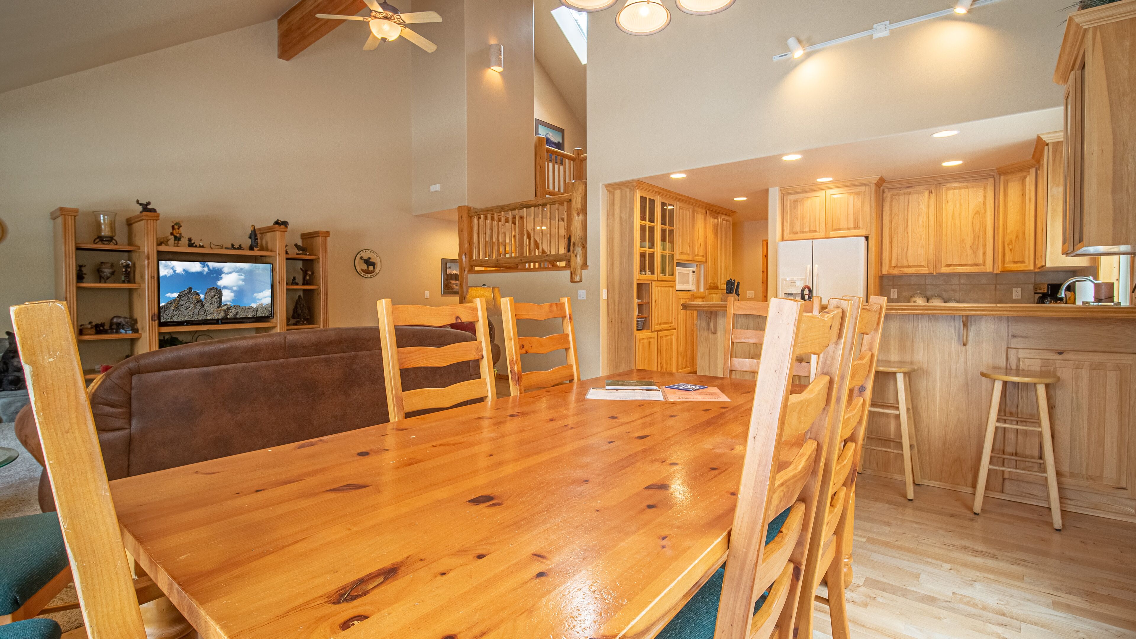 A spacious living room with a wooden dining table, a sofa, and a television. The kitchen features wooden cabinets and a refrigerator.