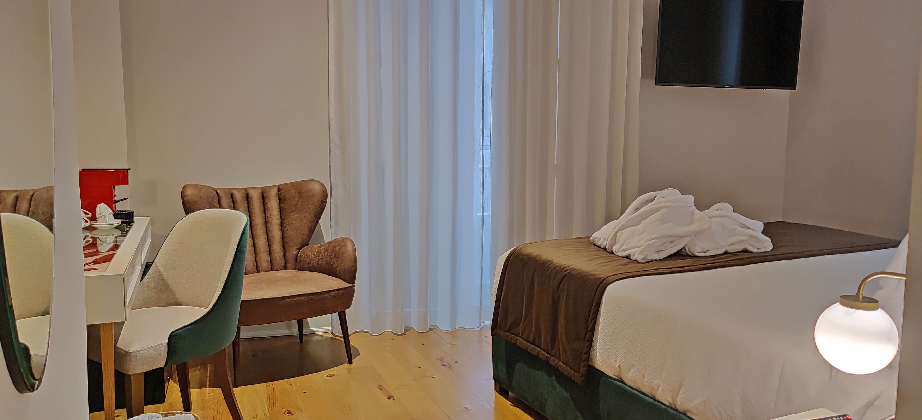 A hotel room with a bed, a chair, a table with a red lamp, and a TV on the wall.