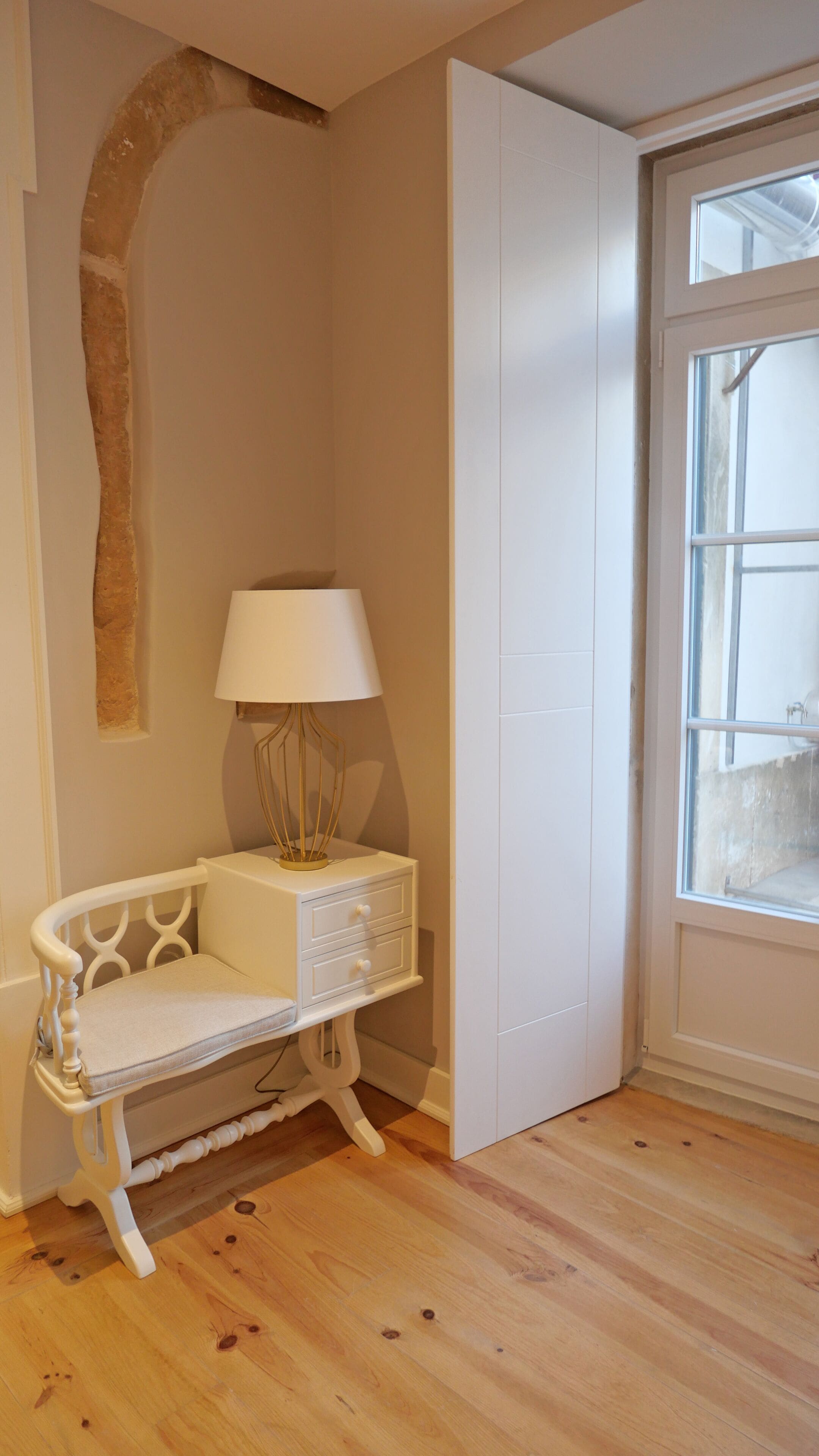 Une chambre d’angle avec une chaise blanche, une petite table de chevet avec une lampe, un plancher en bois et une fenêtre donnant sur un beau paysage.