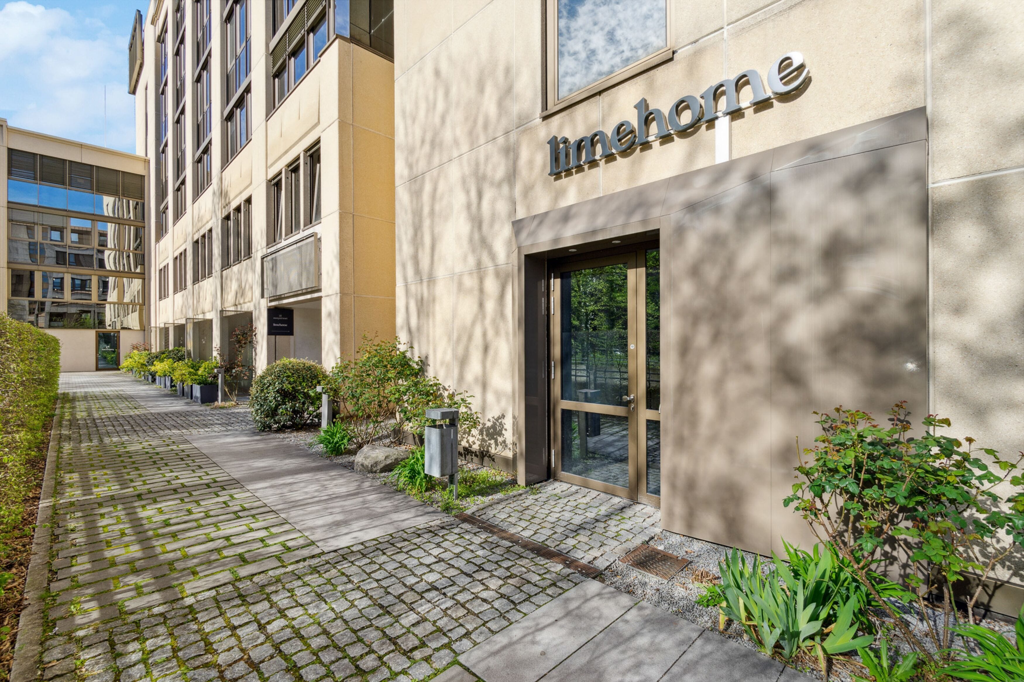 Un edificio moderno con un letrero que dice "limehome". Tiene un camino pavimentado, plantas en macetas y una entrada de vidrio.