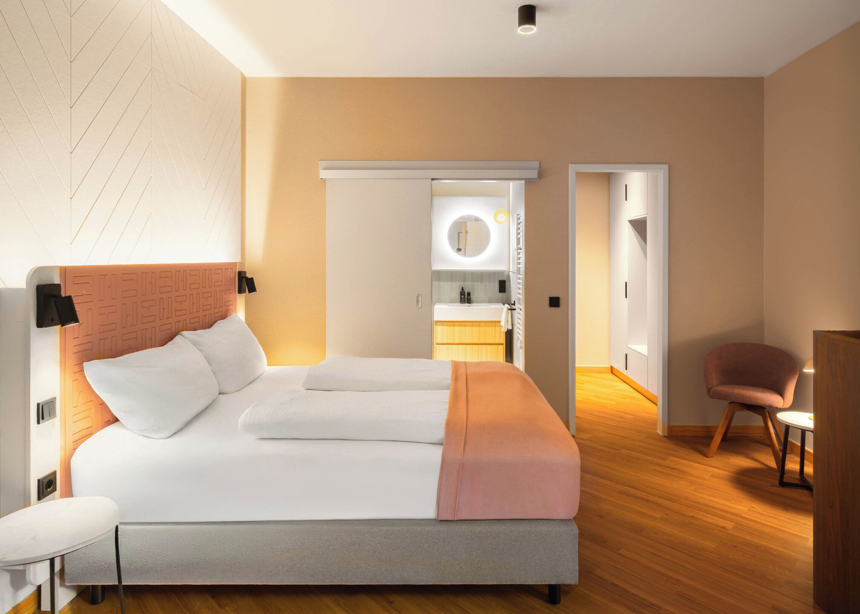 Ein modernes Hotelzimmer mit einem großen Bett, einem Nachttisch, einem Sessel und einem durchgegangenen Tür erkennbaren Badezimmer.