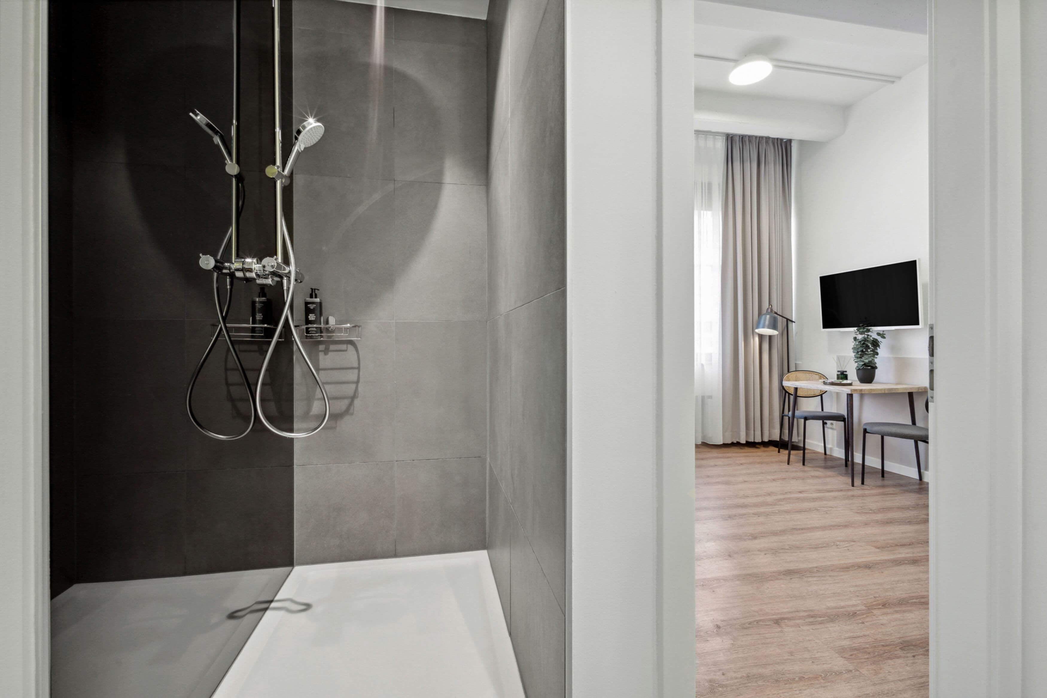 A modern bathroom with a shower, a toilet, and a sink. There is a dining area with a table and chairs visible through an open doorway.