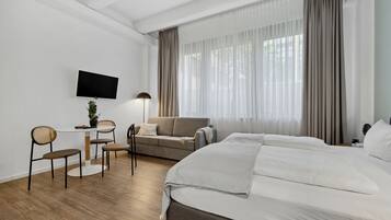 Una habitació d'hotel moderna amb un llit gran, un sofà, un escriptori amb cadires, un televisor fixat a la paret i grans finestres amb cortines.