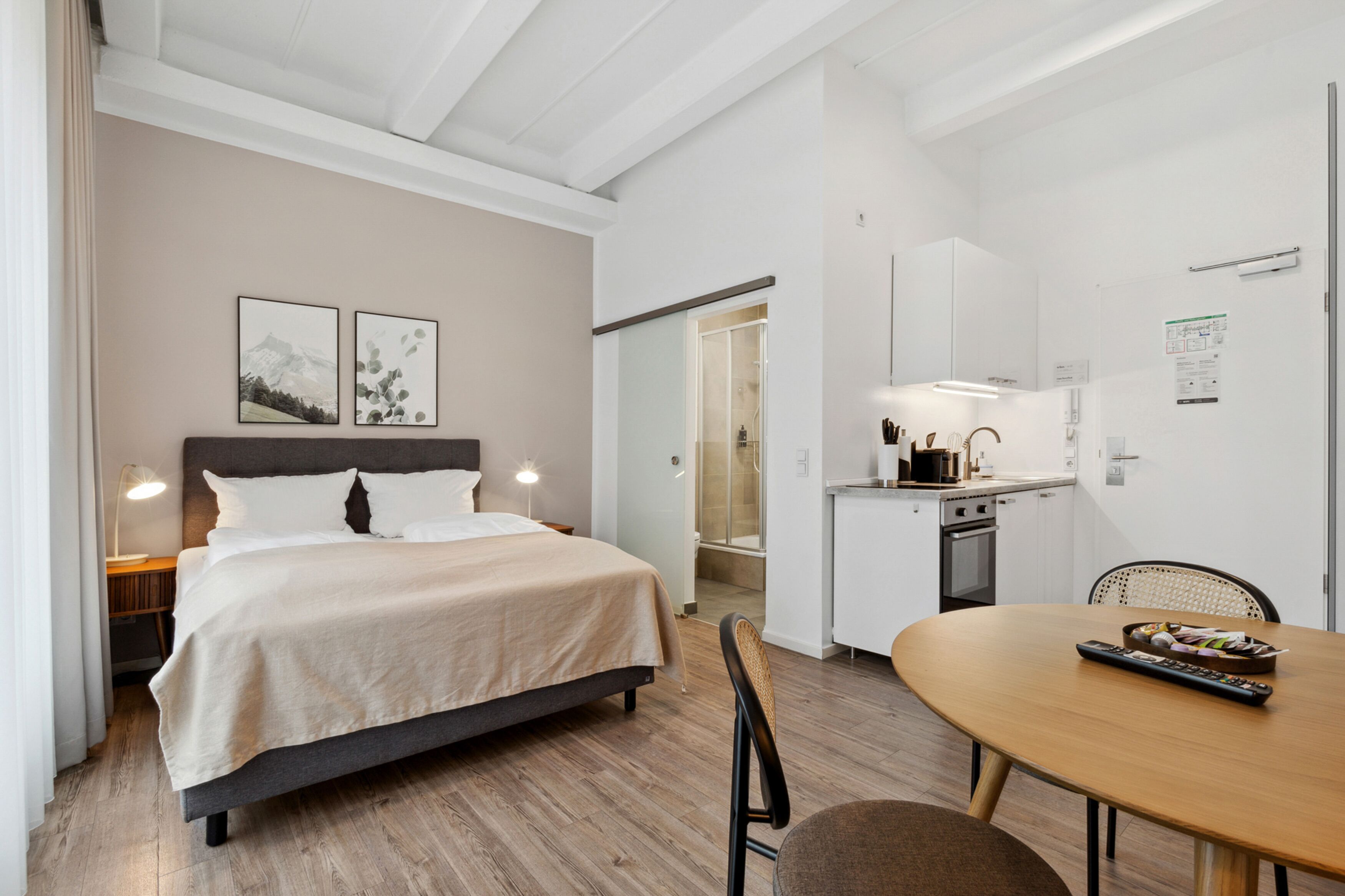 Ett modernt hotellrum med en säng, ett nattduksbord, en lampa och utsikt över badrummet och köket.