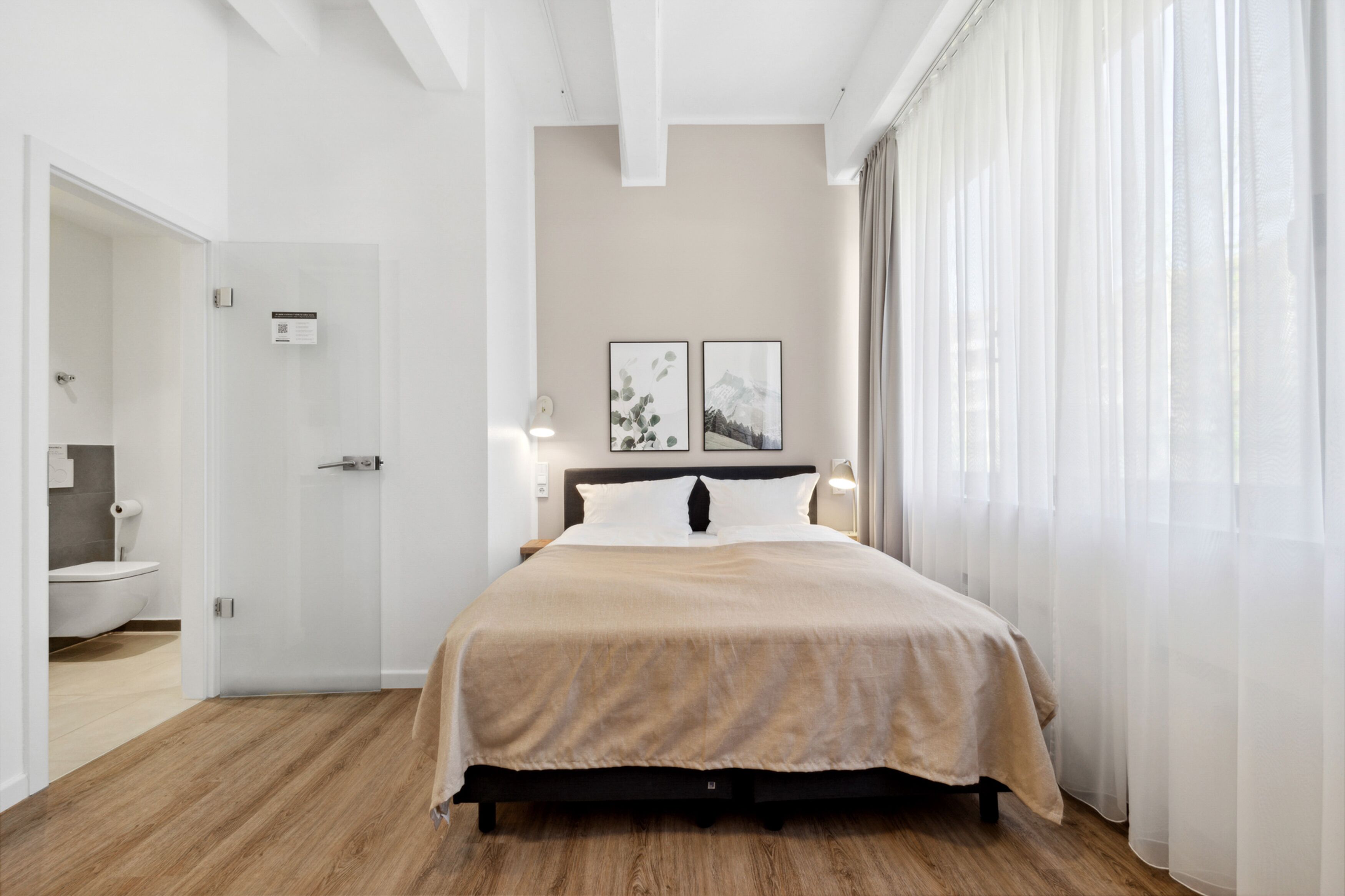 A modern bedroom with a large bed, a bedside table, and a nightstand. There is a bathroom visible to the left with a toilet and sink.