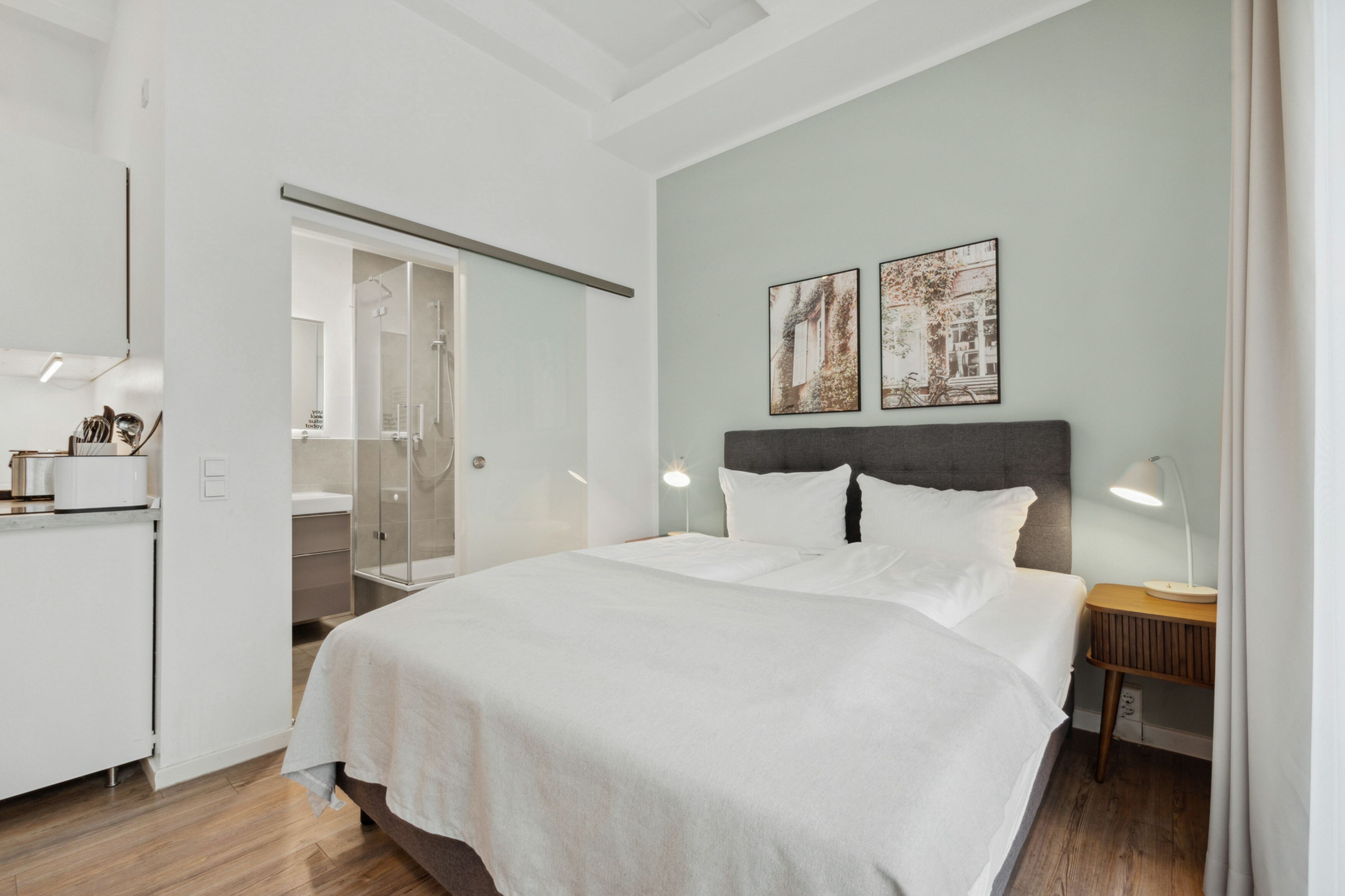 Habitación de hotel moderna con una cama grande, una mesita de noche con lámpara, un armario y un baño visible a través de una puerta de cristal.