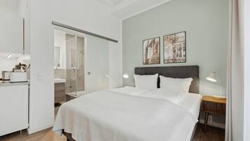 Habitación de hotel moderna con una cama grande, una mesita de noche con lámpara, un armario y un baño visible a través de una puerta de cristal.
