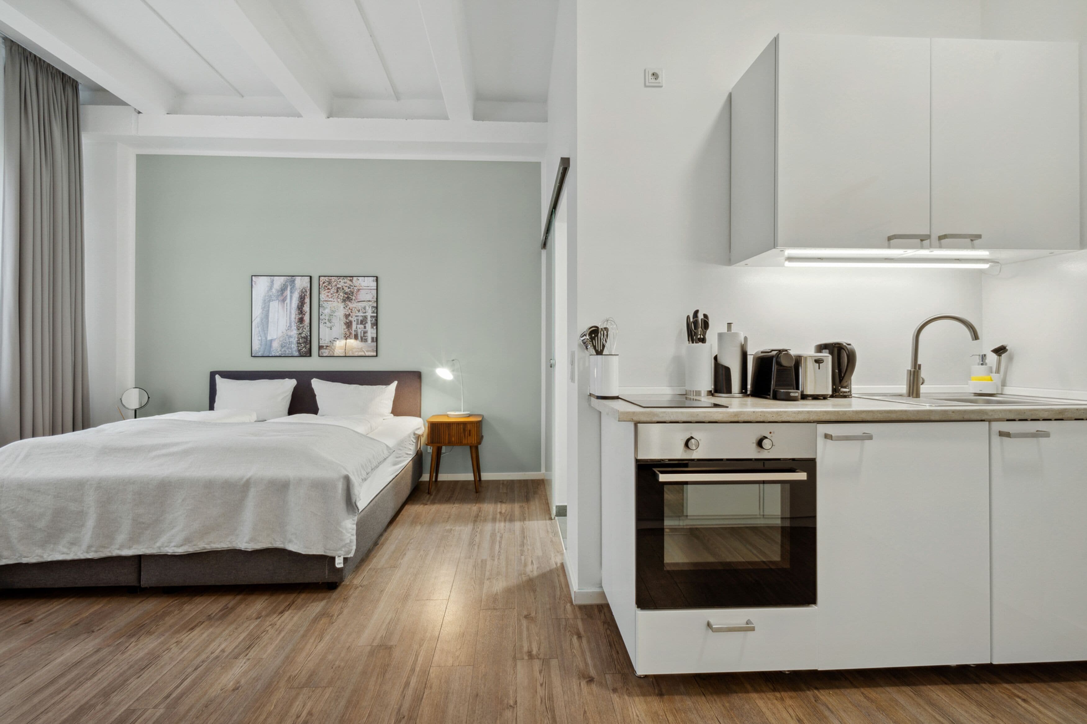 A modern hotel room with a bed, bedside table, lamp, and a kitchenette with a stove and cabinets.
