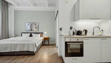 A modern hotel room with a bed, bedside table, lamp, and a kitchenette with a stove and cabinets.