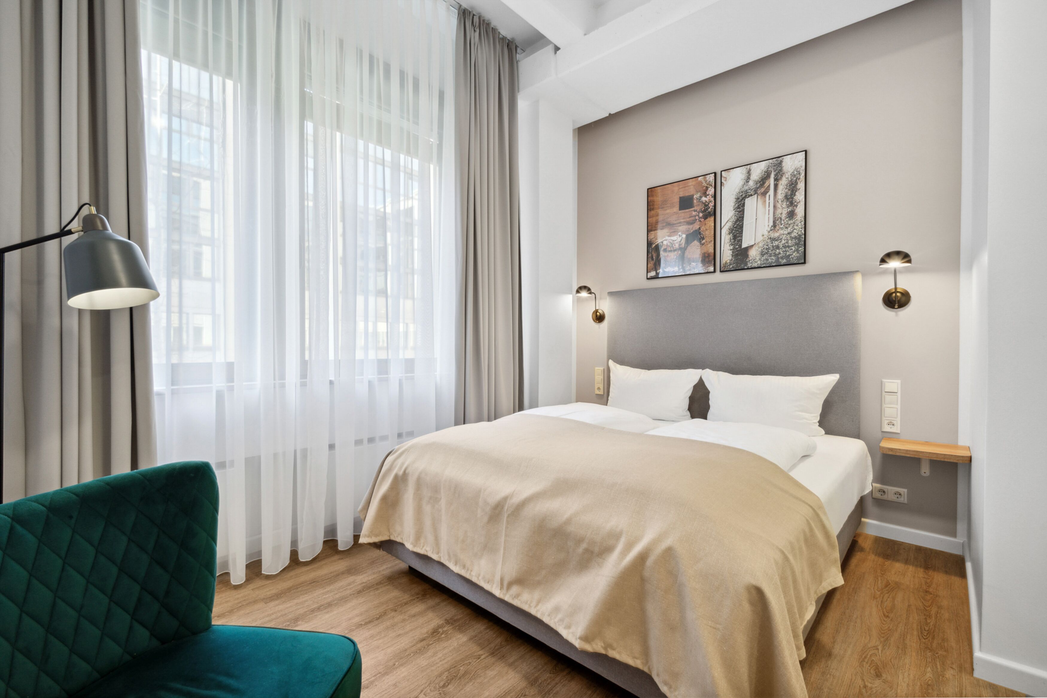 A modern hotel room with a large bed, a green armchair, and a view of the city through sheer curtains.