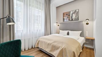 A modern hotel room with a large bed, a green armchair, and a view of the city through sheer curtains.