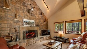 A cozy living room with a stone fireplace, leather furniture, and large windows offering a forest view.