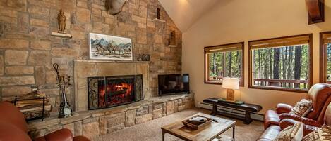 A cozy living room with a stone fireplace, leather furniture, and large windows offering a forest view.