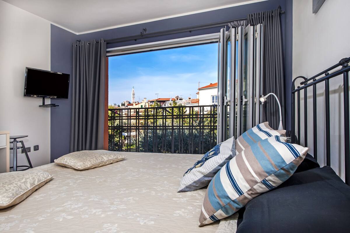 Une chambre à coucher avec un lit, une télévision et une vue sur le paysage urbain, visible depuis un balcon.