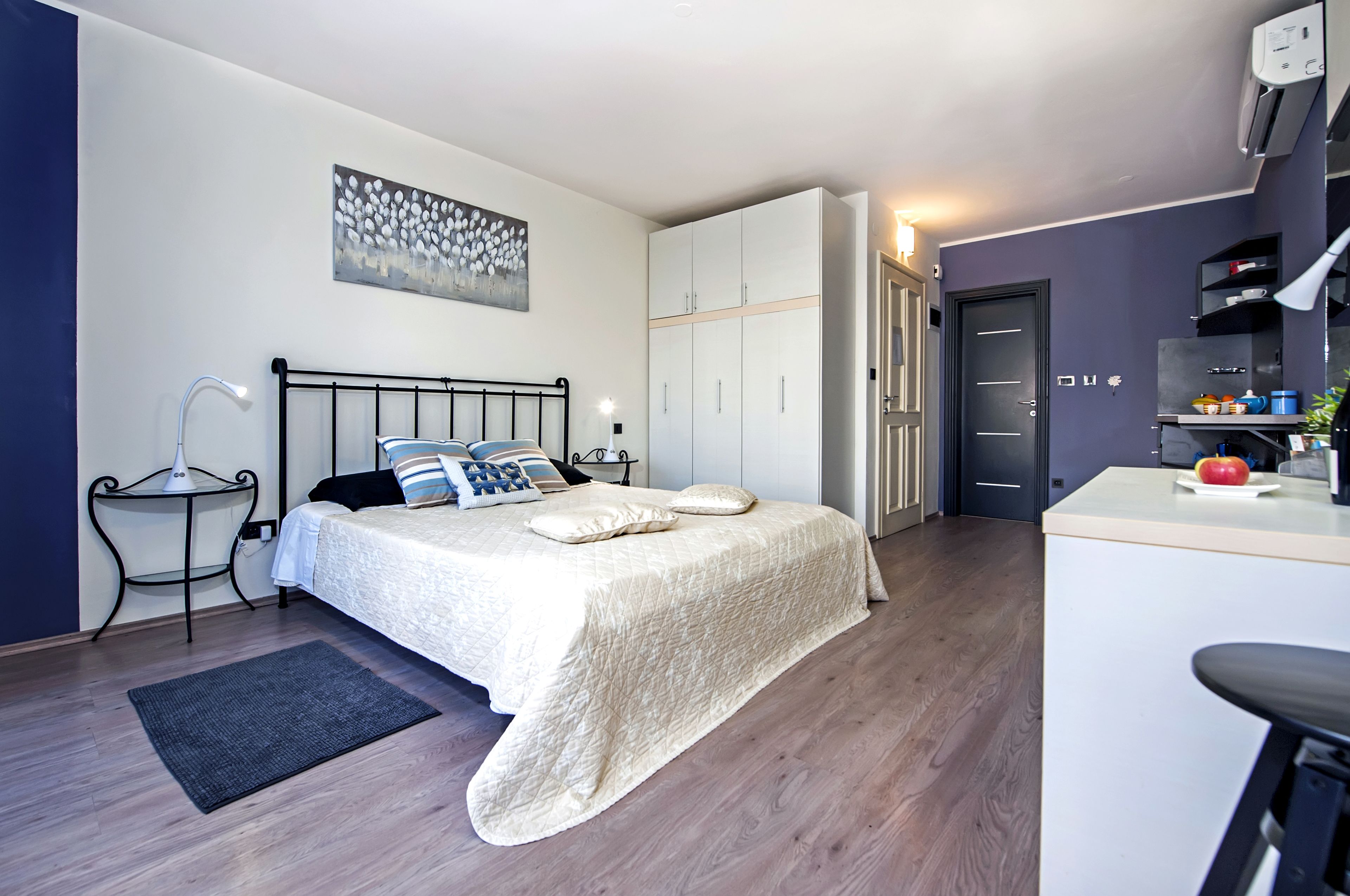 A bedroom with a bed, bedside table, lamp, wardrobe, and a small kitchenette area.