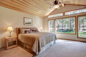 A bedroom with a large bed, two bedside tables with lamps, a ceiling fan, and large windows with a forest view. - Cozy Sunriver Home with Exclusive Golf (Sunriver)