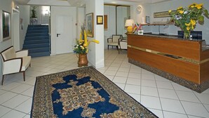 Un hall d’hôtel comprenant un comptoir d’accueil, une moquette bleue, un escalier et un espace d’attente avec des chaises et un vase de fleurs.