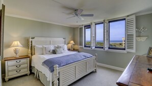 Un dormitorio con cama, mesitas de noche, ventilador de techo y vista al océano a través de contraventanas abiertas.