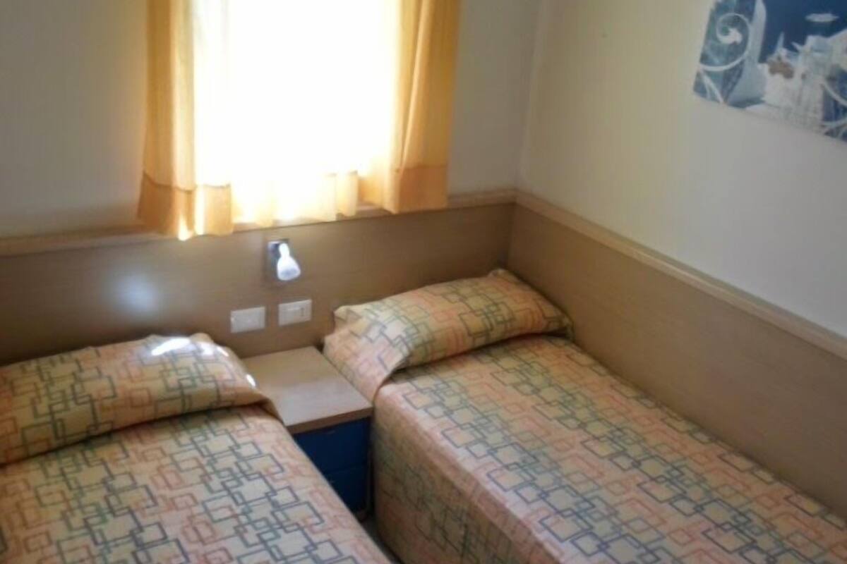 A small room with two single beds, a window with curtains, a wall-mounted lamp, and a poster.