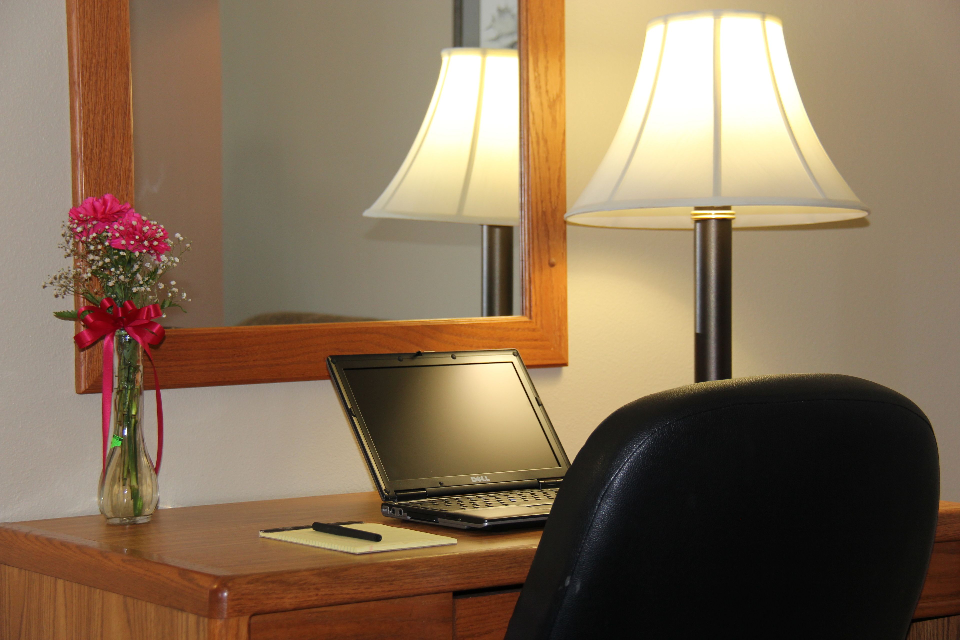 A hotel desk with a laptop, a vase with flowers, a lamp, and a mirror.