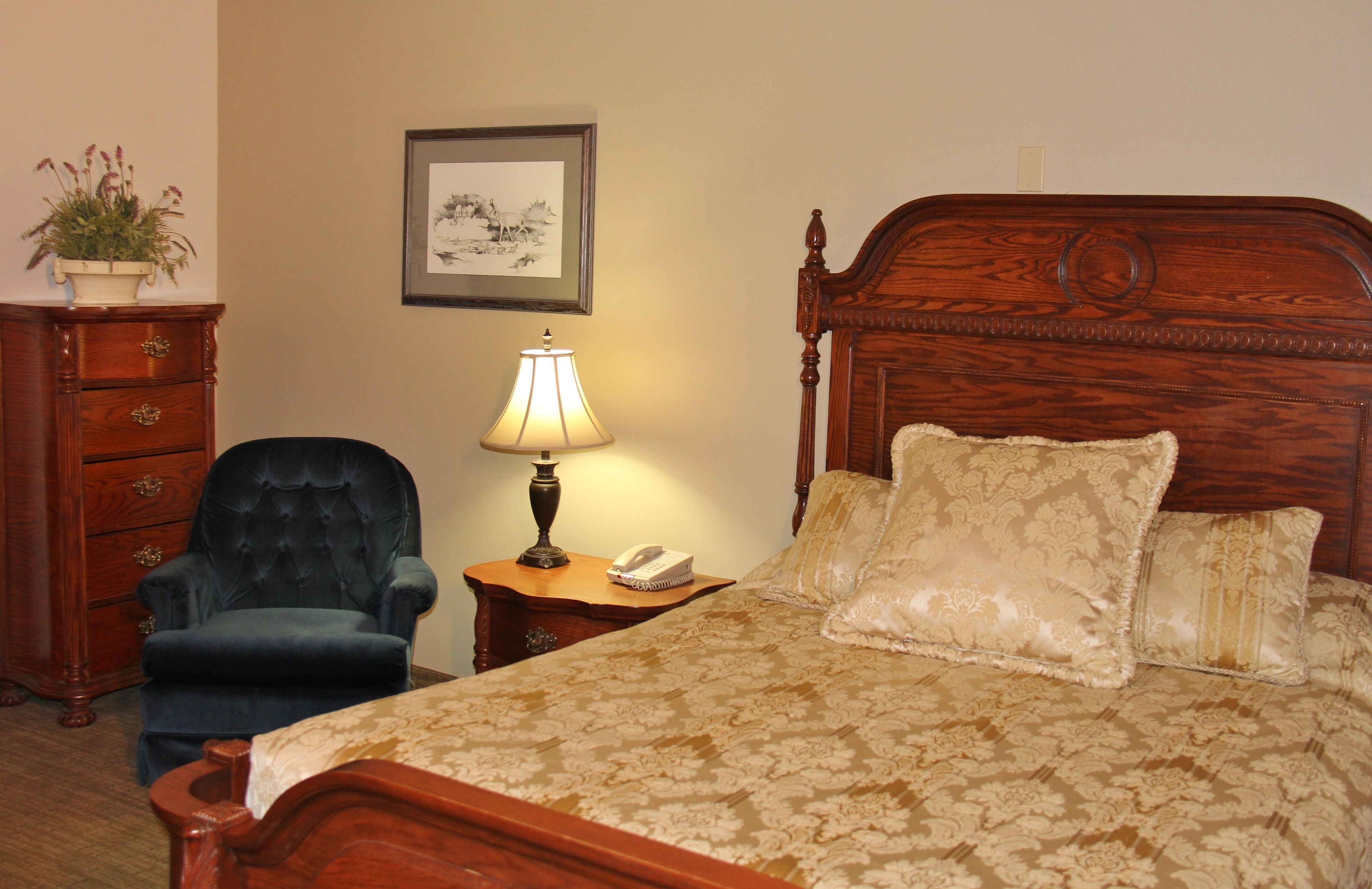 A neatly arranged bedroom with a wooden bed, a dark upholstered chair, a bedside table with a lamp, and a dresser with drawers.