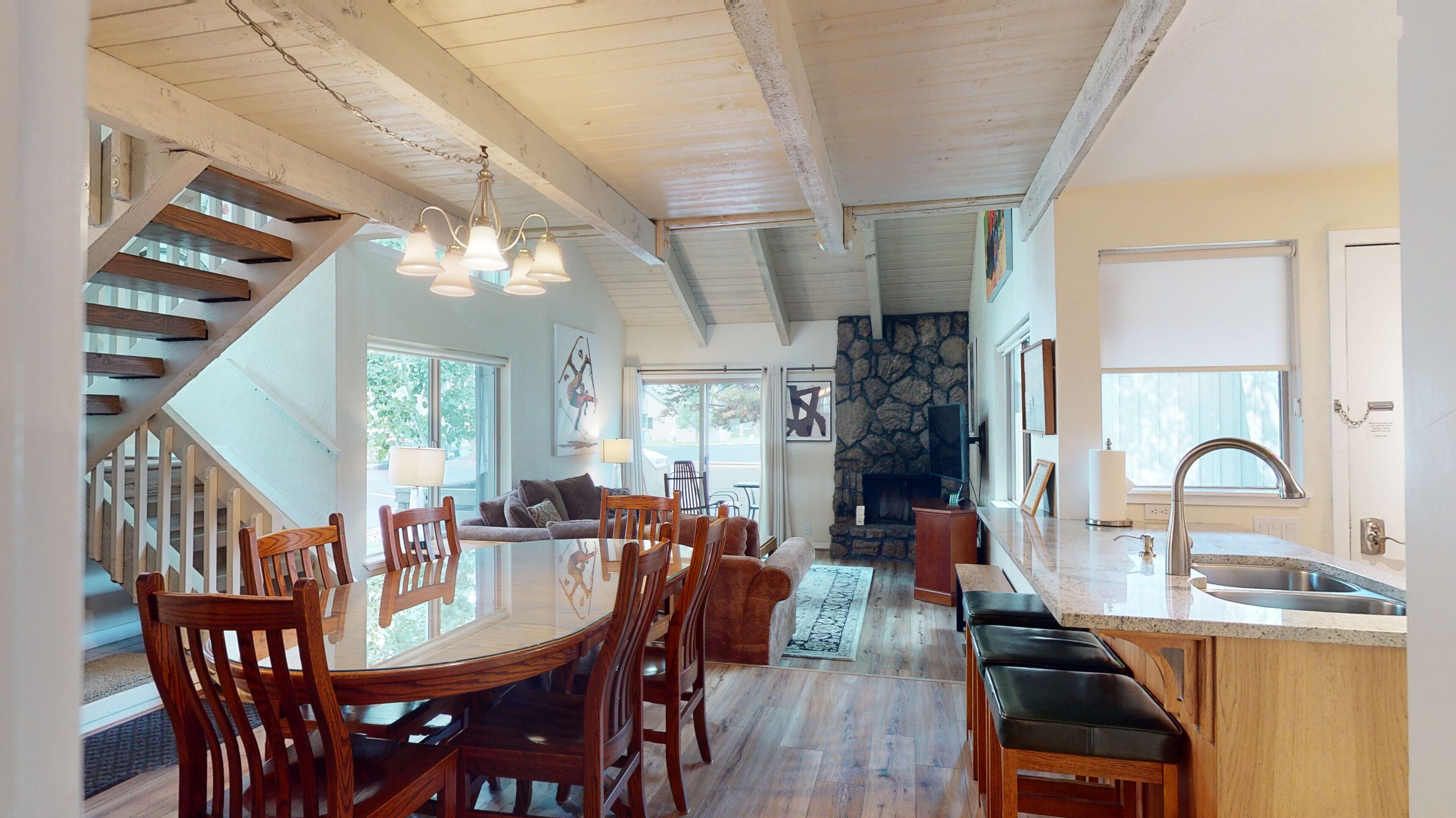 A spacious living area with a wooden staircase, a dining table with chairs, a stone fireplace, and a kitchen with a sink and countertop.