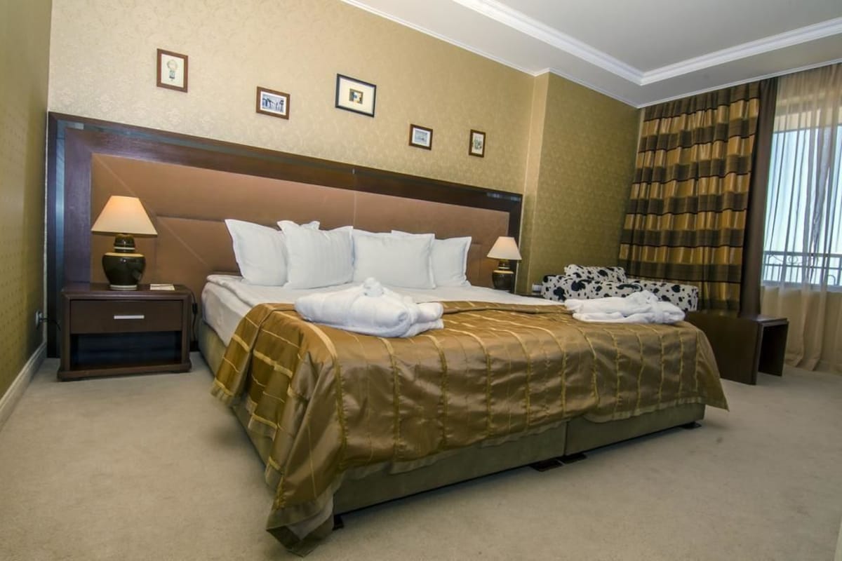 Quarto de hotel com uma cama grande, duas mesas de cabeceira, um painel de cabeceira e uma janela com cortinas.