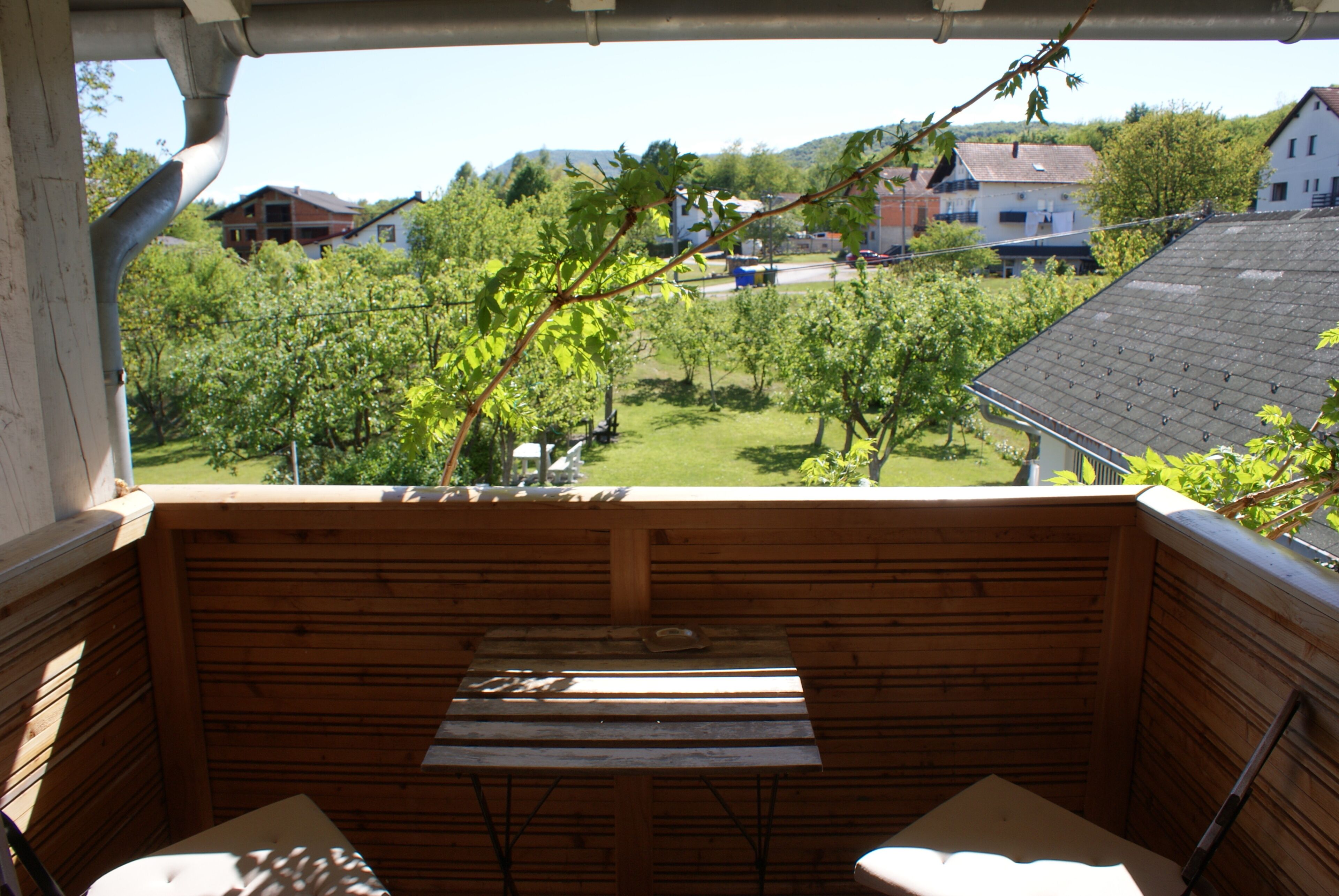 Una terraza de madera con una mesa y sillas, con vistas a un jardín con árboles y casas al fondo.