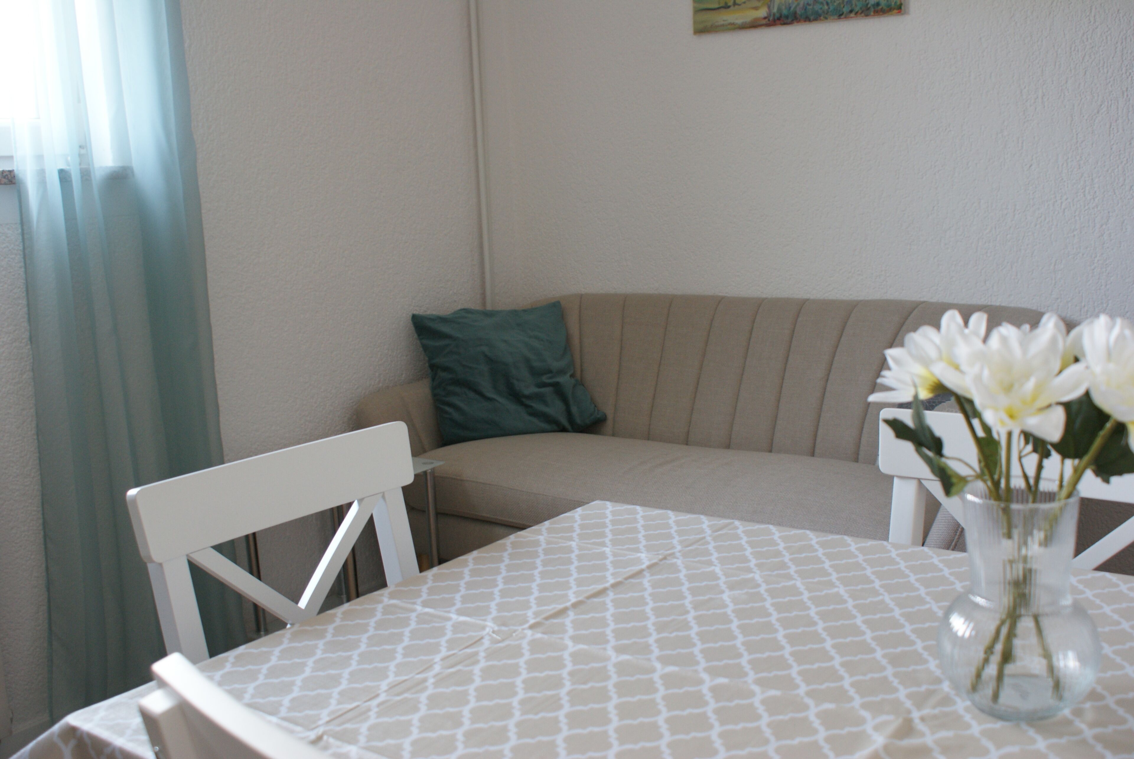 Una habitación pequeña y acogedora con una mesa, una silla, un sofá y un jarrón con flores.