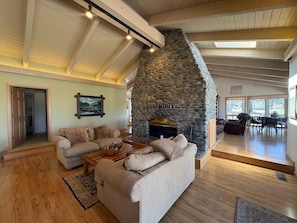 A spacious living room with a stone fireplace, wooden ceiling beams, and a large window providing natural light.