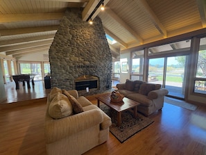 A spacious living area with a stone fireplace, wooden ceiling, and large windows offering a view of the outdoors.