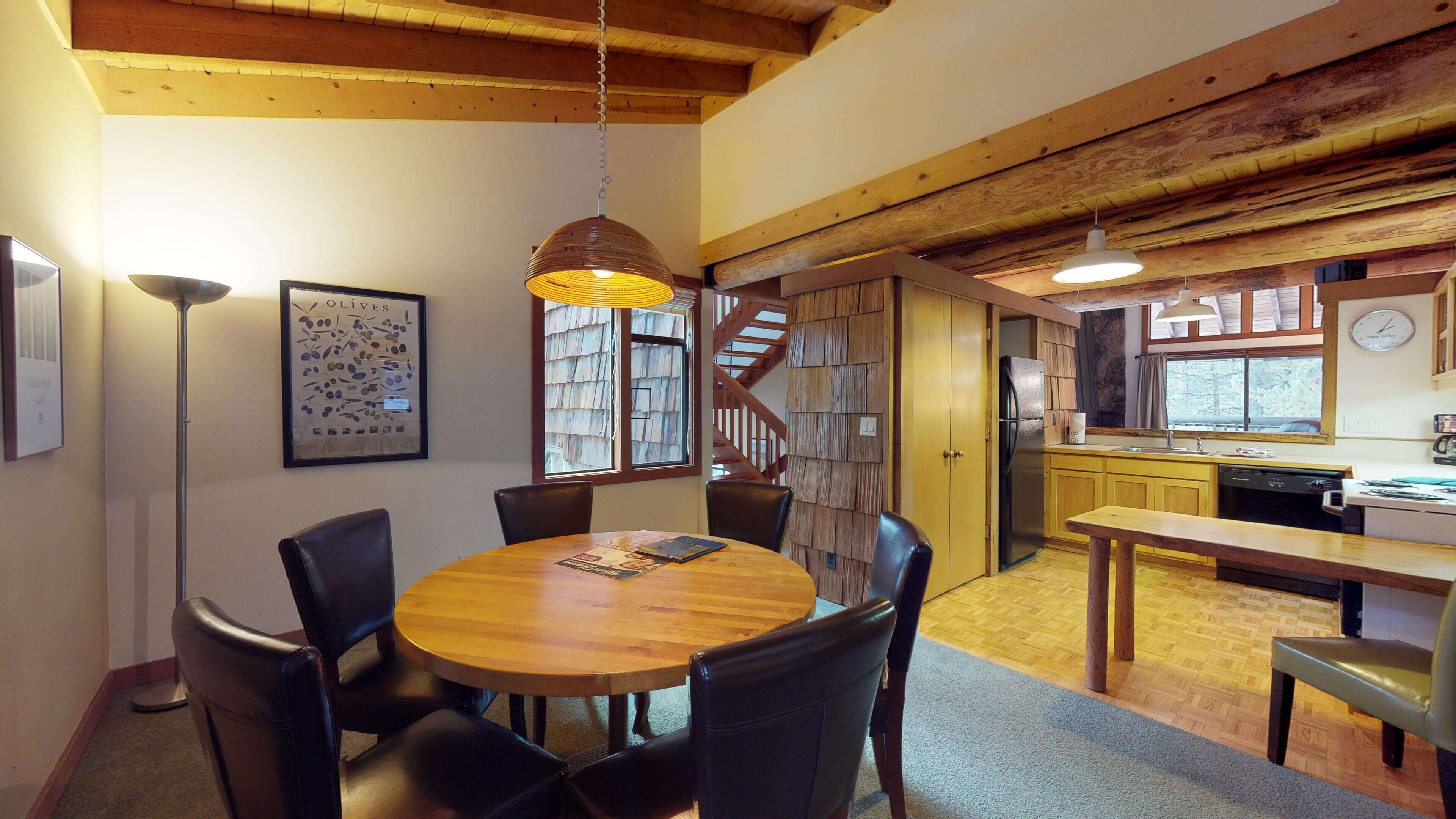 Un espace repas avec une table et des chaises en bois, un luminaire suspendu et un escalier menant à une cuisine.