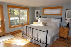 Een slaapkamer met een groot raam, een bed met metalen frame, houten nachtkastjes en een houten vloer.