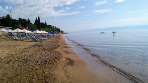 Ein Strand mit Liegestühlen und Sonnenschirmen, ruhigem Meer und hügeliger Landschaft im Hintergrund.