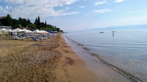Ein Strand mit Liegestühlen und Sonnenschirmen, ruhigem Meer und hügeliger Landschaft im Hintergrund.