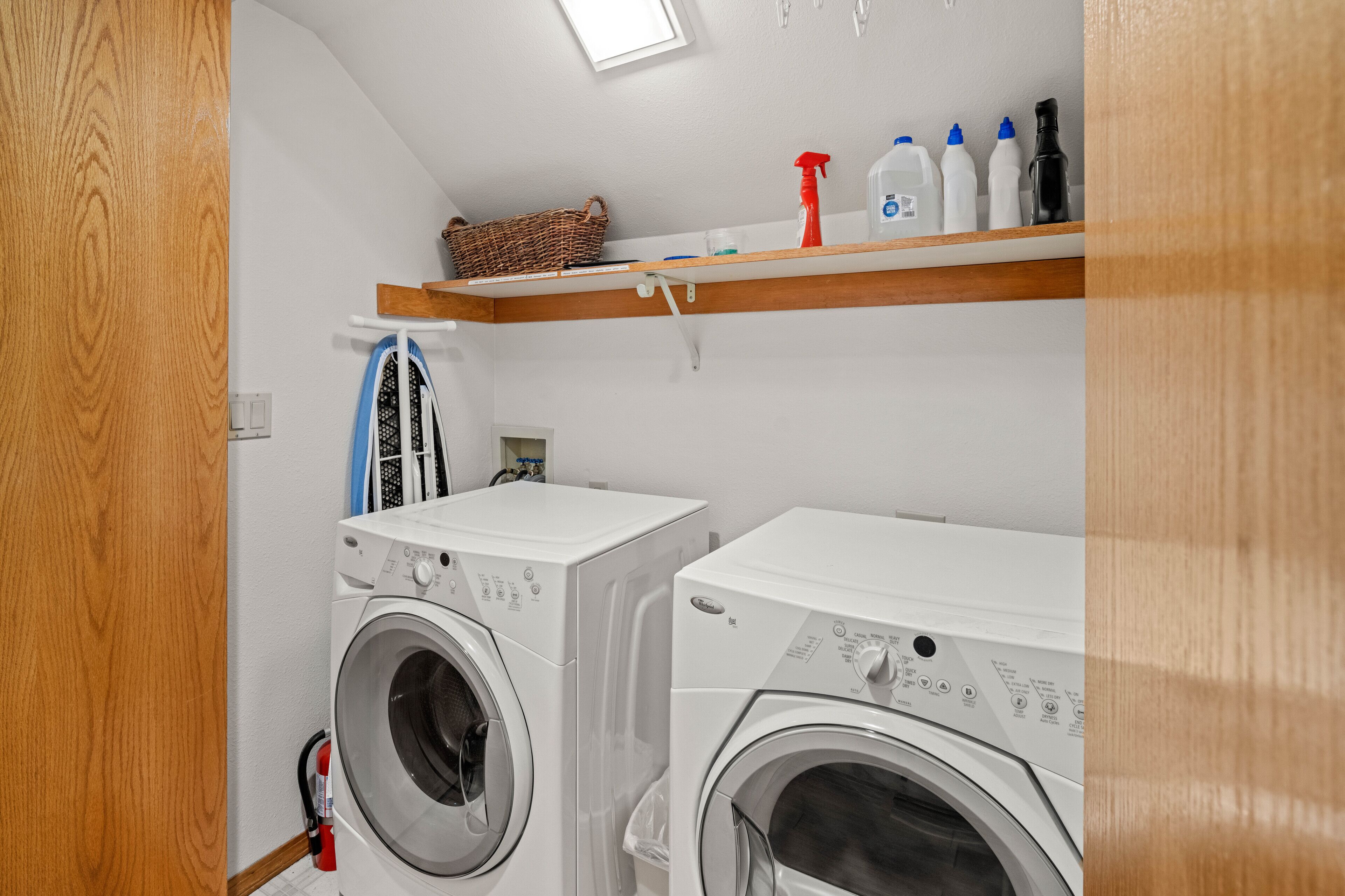 A laundry room with two front-loading washing machines, a shelf with cleaning supplies, and a basket.