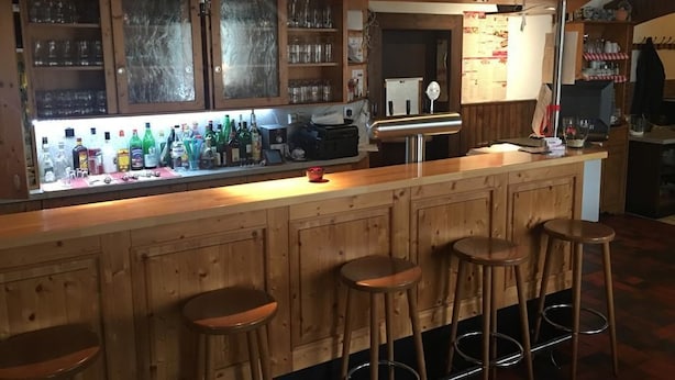 A bar area with wooden stools, a counter with various bottles, and shelves with glasses.