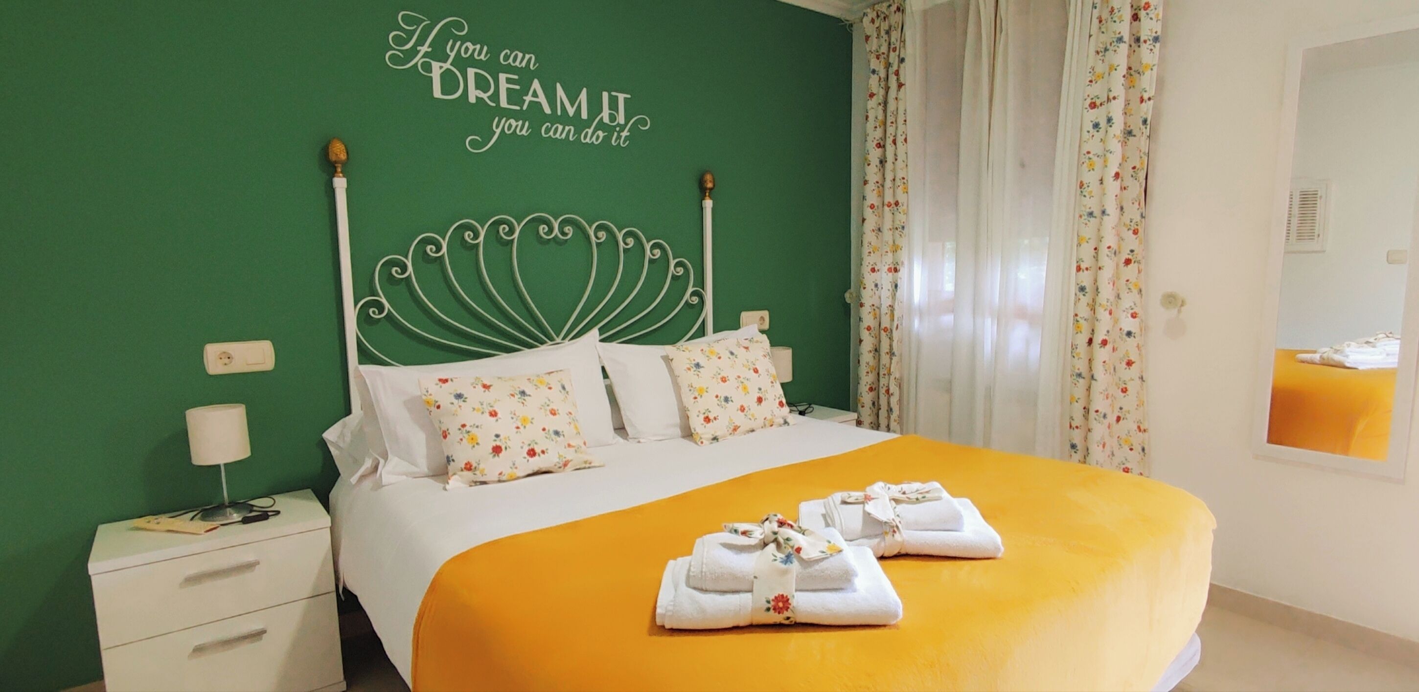A neatly made bed with floral pillows, a green wall with a motivational quote, and a yellow bedspread.