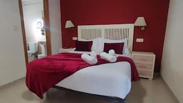 Un lit bien fait, avec des draps blancs et une couverture rouge, deux oreillers blancs et deux serviettes enroulées posées sur le lit.