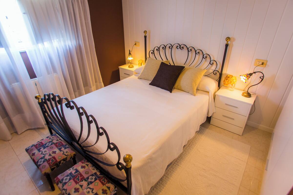 A neatly made bed with a headboard, two bedside tables with lamps, and a bench with a floral pattern.