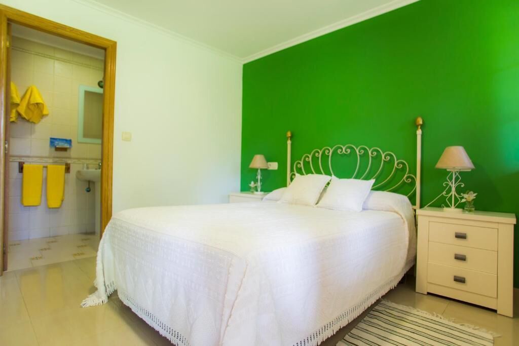 Un dormitorio con pared verde, cama blanca con cabecera decorativa, dos mesitas de noche con lámparas y un baño visible a través de una puerta abierta.