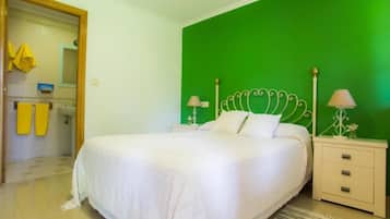 Un dormitorio con pared verde, cama blanca con cabecera decorativa, dos mesitas de noche con lámparas y un baño visible a través de una puerta abierta.
