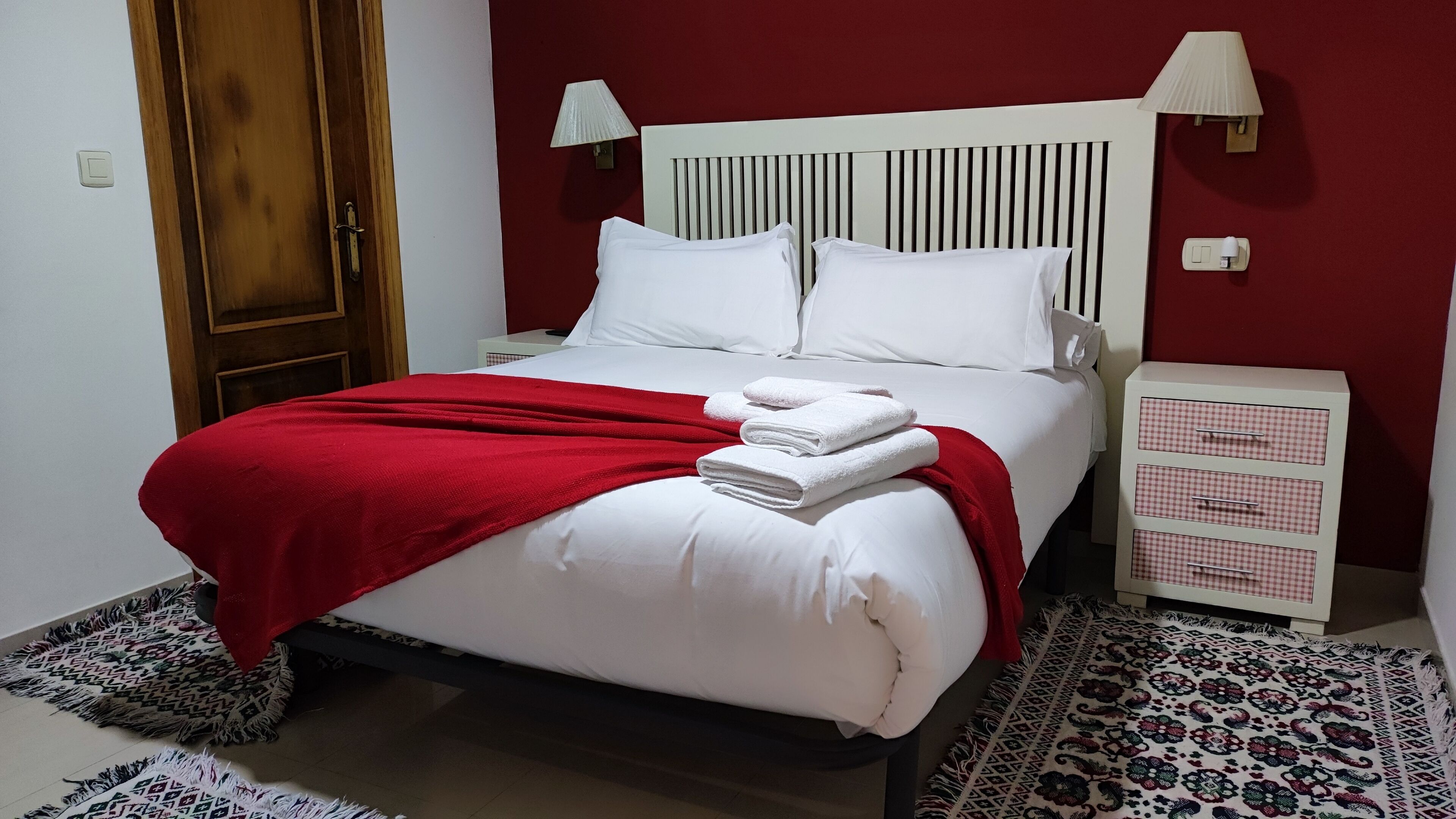 Een net opgemaakt bed met witte lakens en een rode deken, een nachtkastje met een lamp, en een kamerbreed tapijt op de vloer.