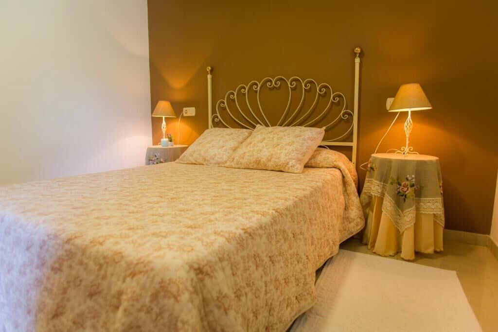 Cama bem arrumada com colcha estampada, duas luminárias de cabeceira e uma mesa decorativa com toalha de mesa florida.