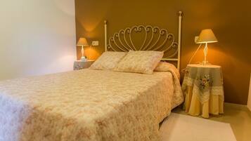 Cama bem arrumada com colcha estampada, duas luminárias de cabeceira e uma mesa decorativa com toalha de mesa florida.