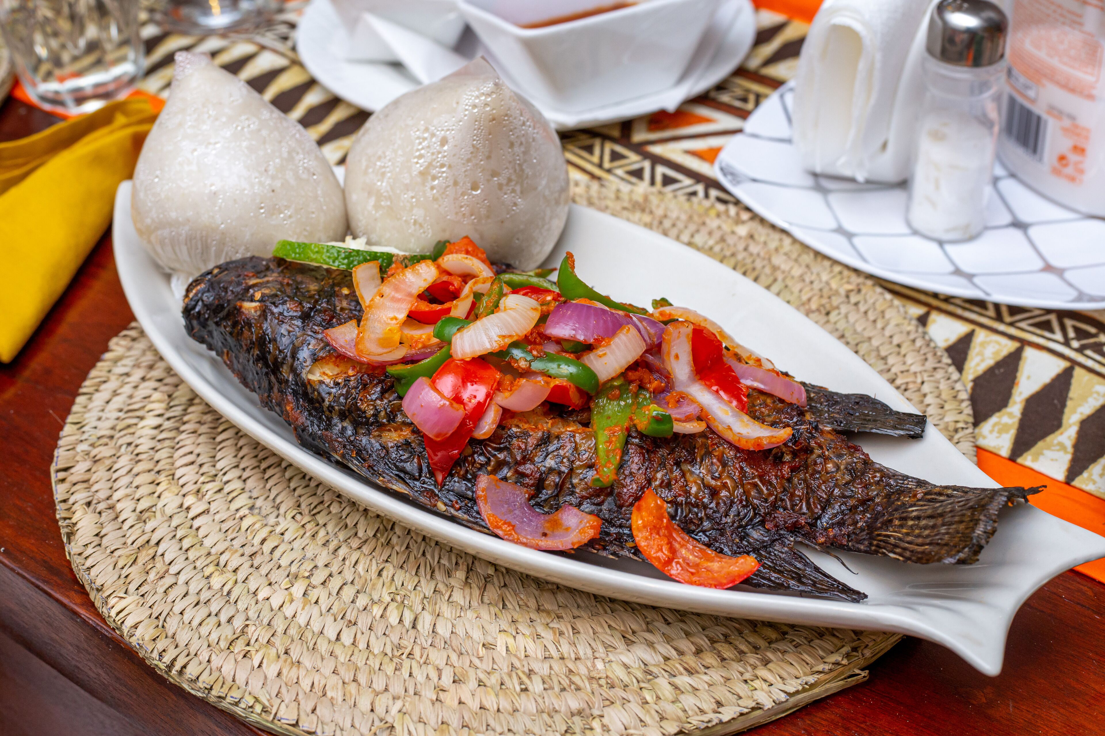 A plate of grilled fish with vegetables and two steamed buns on a woven placemat.