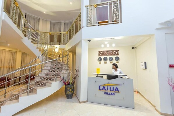 A hotel reception area with a receptionist desk, a staircase with patterned railings, and a decorative wall with clocks. - La Tua Villas (Prampram)