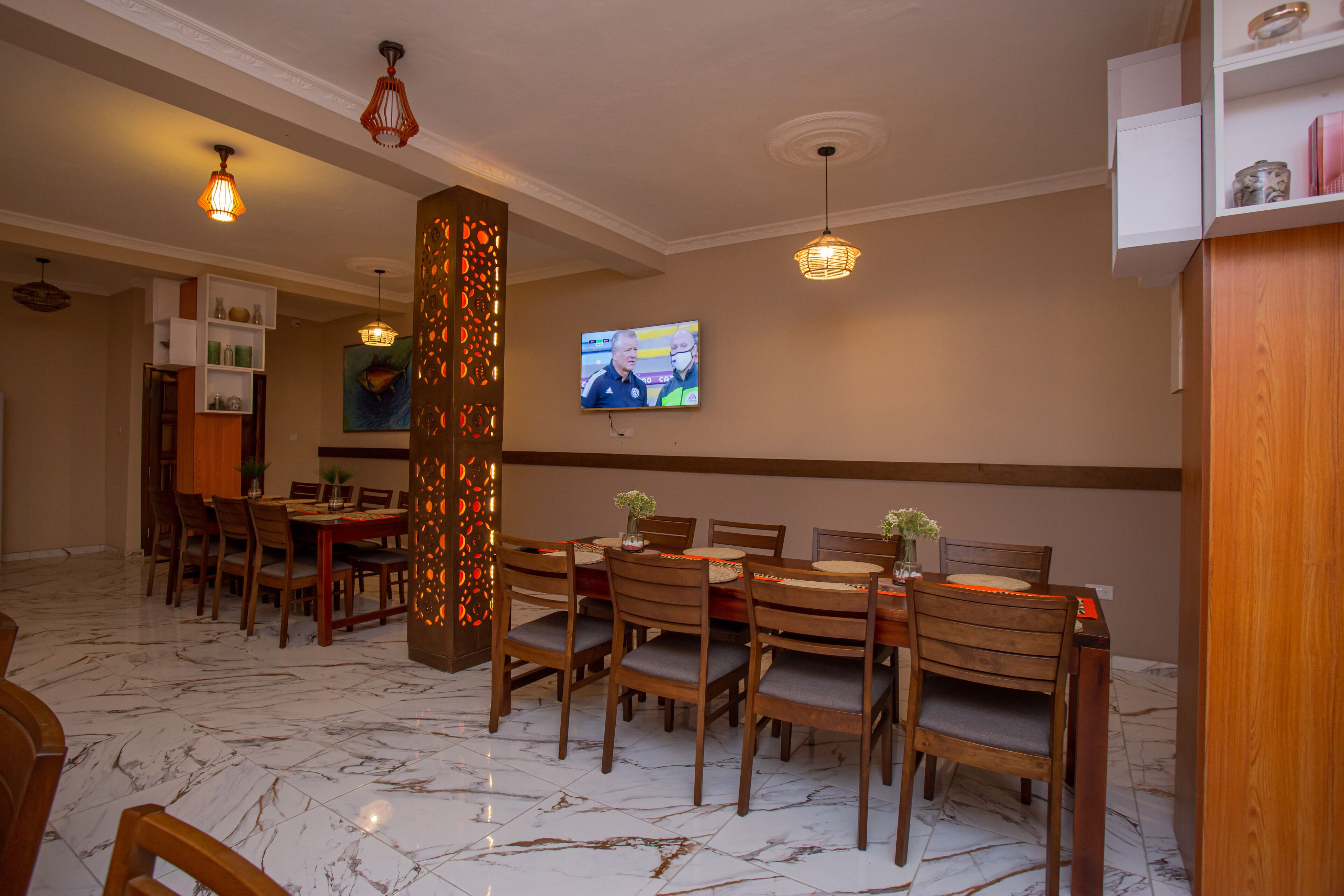 A dining area with wooden tables and chairs, a television mounted on the wall, and a decorative wall panel.
