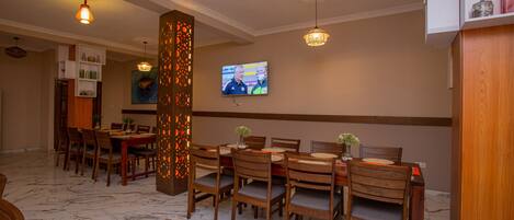 A dining area with wooden tables and chairs, a television mounted on the wall, and a decorative wall panel.