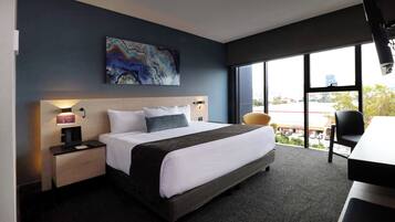 A modern hotel room with a large bed, a desk, and a chair. There is a painting on the wall and a window with a view of the city.